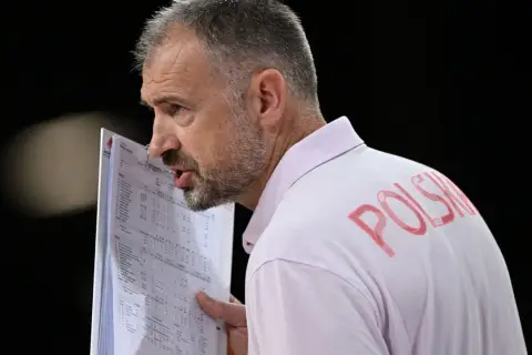 Trener Nikola Grbić wpatrzony w dokumenty, z napisem "POLSKA" na koszulce. Może analizuje statystyki, myśląc o swojej żonie.