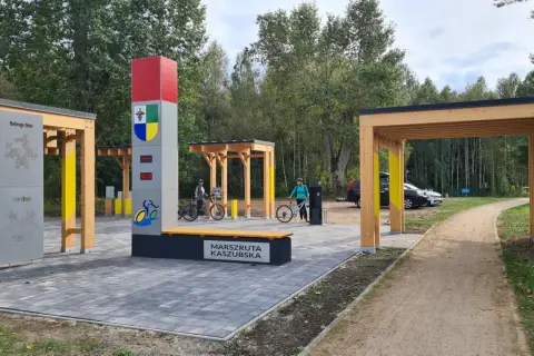 Nowiutka stacja rowerowa na Marszrucie Kaszubskiej. Dwie panie z rowerami, nowoczesna infrastruktura i leśne otoczenie.