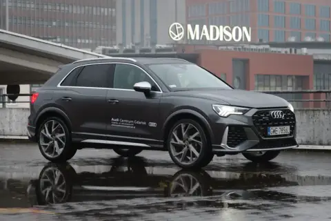 Szary Audi RS Q3 na mokrej nawierzchni, z odbiciem w kałuży. W tle budynek z napisem MADISON.