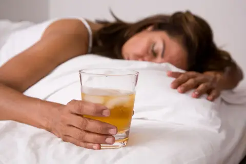 Kobieta śpi w łóżku, trzymając szklankę z napojem i lodem. Może to być alkohol, np. GHB.