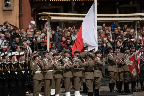 Orkiestra wojskowa grająca na instrumentach dętych na tle tłumu i polskiej flagi.