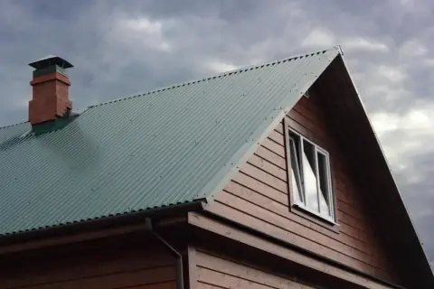 Dach domu pokryty zieloną blachą trapezową, widoczny komin i okno. Szerokość krycia blachy trapezowej zapewnia szczelność.
