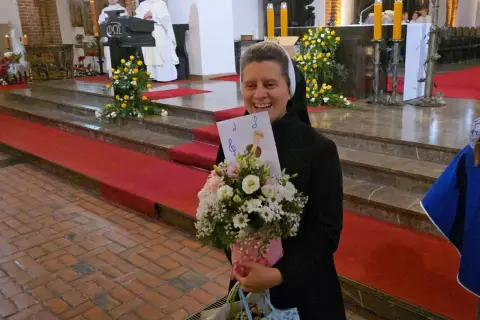 Radosna zakonnica z bukietem kwiatów i kartką z nutami. W tle msza święta, 25 października.