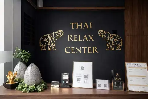Recepcja centrum masażu tajskiego. Złote słonie i napis "THAI RELAX CENTER" na czarnej ścianie. Na blacie dekoracje i cennik. Tu możesz się wyciszyć.