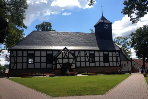 Kościół z wieżą zegarową, otoczony zielenią i ławkami. W tle niebo z chmurami. Miejsce pielgrzymek do Matki Boskiej Sianowskiej.