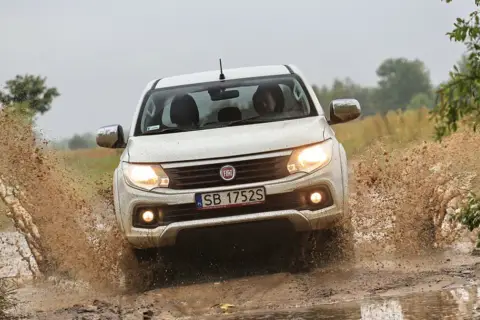 Biały Fiat Fullback z impetem wjeżdża w błotnistą kałużę, wzbijając fontanny brudu.