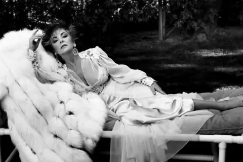 Kobieta w białym futrze i jedwabnej sukni, w stylu Helmut Newton, leży na leżance w ogrodzie.