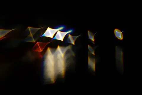 Rozmyte kształty z tęczowymi obwódkami, przypominające gwiazdy i trójkąty, unoszą się na czarnym tle. Widoczna jest wyraźna aberracja chromatyczna.