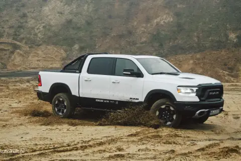 Biały Dodge Ram 1500 z czarnymi dodatkami pokonuje piaszczysty teren, wzbijając błoto.