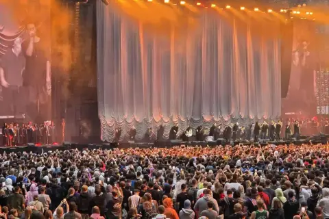 Ogromny tłum na quebo koncercie, fani nagrywają występ na swoich telefonach. Na scenie artyści i muzycy.