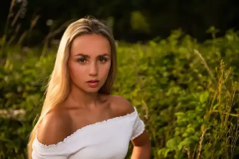Piękna fotografia portretowa młodej kobiety z długimi blond włosami, w białej bluzce, na tle zielonej natury.