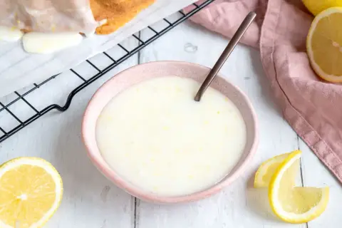 Zitronenglasur selber machen: cremige Glasur in einer Schale, daneben Zitronenspalten und ein Stück Kuchen auf einem Gitter.