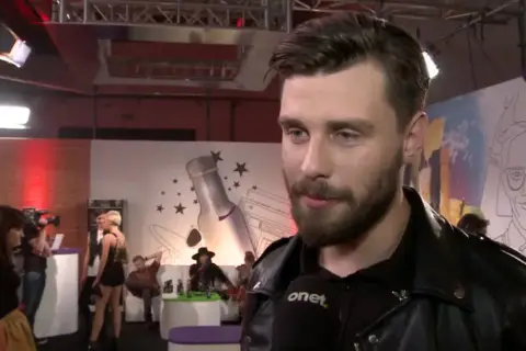 Ernest Staniaszek udziela wywiadu na tle scenografii z butelką i gwiazdami.