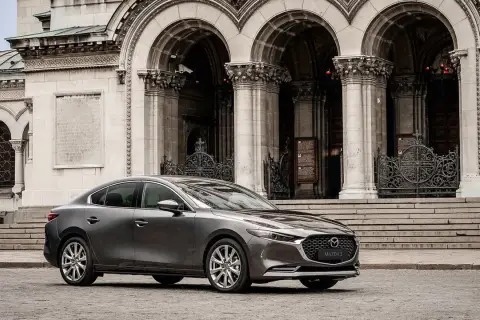 Elegancka, szara Mazda 3 2025 przed monumentalną, kamienną budowlą z arkadami i ozdobnymi bramami.