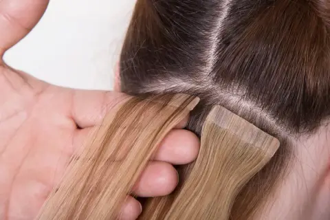 Manos aplicando extensiones adhesivas rubias a cabello castaño. El proceso de colocación de extensiones adhesivas es delicado.
