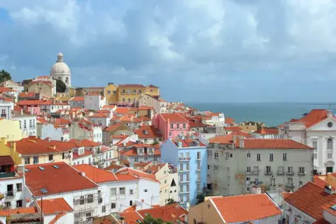 One day in Lisbon, a panorama of colorful buildings with red-tiled roofs stretches towards the shimmering blue sea under a cloudy sky.