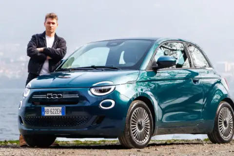 Młody mężczyzna stoi obok swojego nowego, turkusowego fiat mini 500 nad jeziorem.
