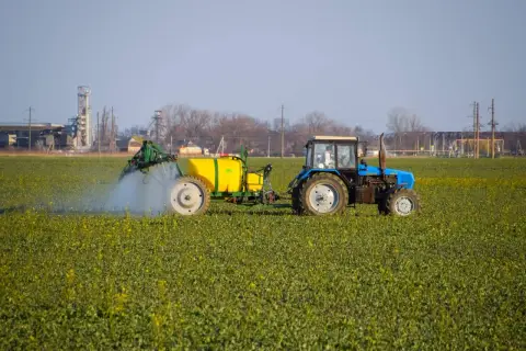 Niebieski traktor z żółtym zbiornikiem wykonuje oprysk na polu zielonych roślin. W tle widać przemysłowe budynki.