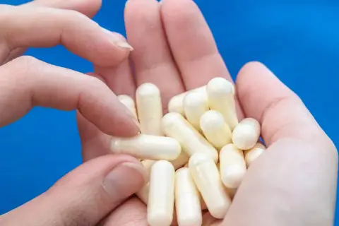 Dłonie trzymają garść białych kapsułek, które mogą zawierać biotynę, wspierającą zdrowie włosów i paznokci.