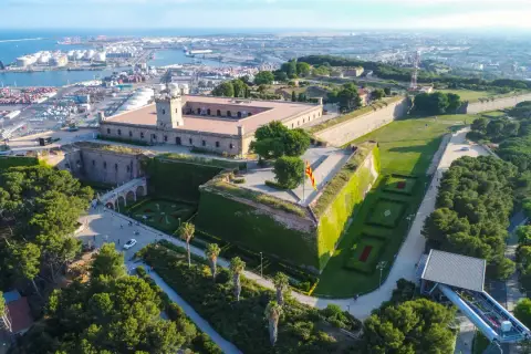 Zamek Castell de Montjuïc góruje nad portem w Barcelonie. Otoczony zielenią, z flagą Katalonii powiewającą na wietrze.