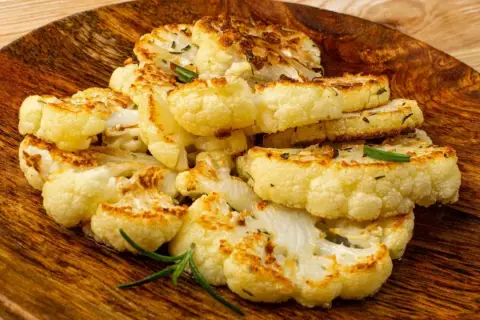 Tranches de chou-fleur rôti, dorées et garnies d'herbes, servies sur un plat en bois.