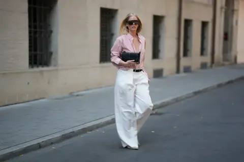 Kobieta w stylowym, różowo-białym paszku Massimo Dutti, szerokich białych spodniach i okularach przeciwsłonecznych idzie ulicą z czarną kopertówką.