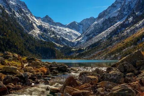 Majestuosas montañas nevadas flanquean el sereno lac de Gaube, con un arroyo rocoso que desemboca en sus aguas turquesas.