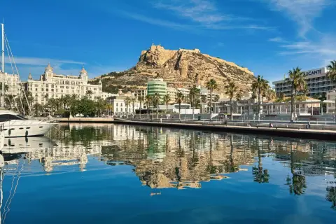 Alicante, luty. Słoneczny dzień nad zatoką z odbiciem zamku Santa Barbara i palm w krystalicznie czystej wodzie.