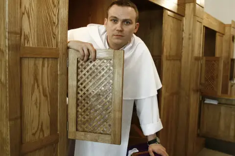 Młody zakonnik w białej sutannie otwiera kratkę konfesjonału, gotów wysłuchać spowiedzi.