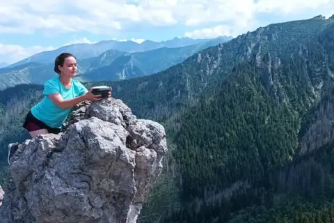 Kobieta na skale z urządzeniem w rękach, podziwia rozległą panoramę górską. Widok zapiera dech w piersiach, to prawdziwa przełęcz w tatrach wysokich.