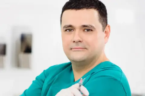 Lekarz w zielonym fartuchu trzyma strzykawkę. W tle widać elementy gabinetu. Wiele pozytywnych klinika ambroziak opinie potwierdza jego profesjonalizm.