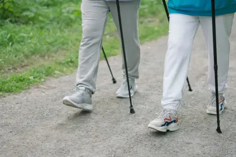 Dwie osoby uprawiające **nordic walking** na żwirowej ścieżce, otoczone zielenią.