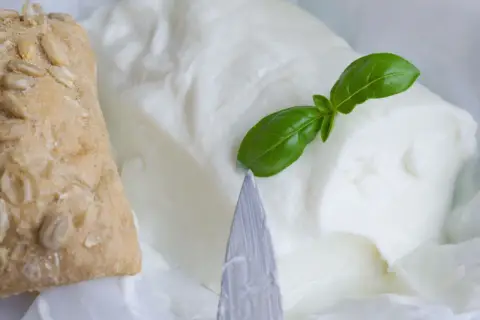 Un panino integrale con semi e un blocco di cremoso stracchino, un formaggio magro, guarnito con una foglia di basilico.
