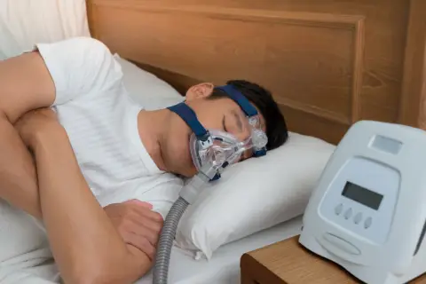 Mann schläft mit CPAP-Maske, um zu wenig Sauerstoff im Schlaf Symptome zu behandeln. Ein Gerät steht daneben.