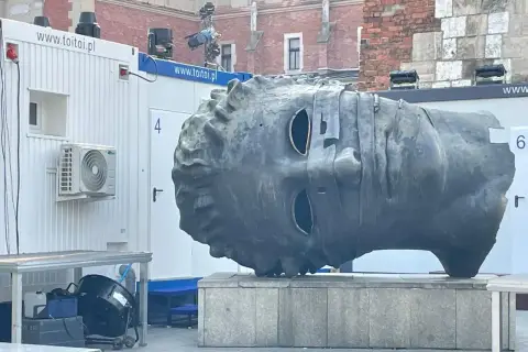 Monumentalna głowa na Rynku w Krakowie, z pustymi oczodołami, stoi obok białych kontenerów.