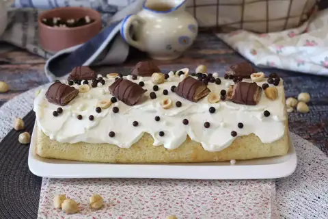 Puszysty biszkopt z kremem, udekorowany kawałkami batoników Kinder Bueno White, orzechami i czekoladowymi kuleczkami.