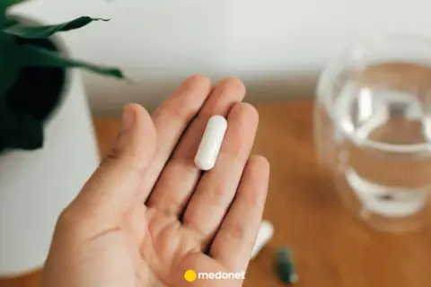 Dłoń trzyma białą kapsułkę. Obok szklanka wody i roślina. Uważaj na nadmiar magnezu objawy.