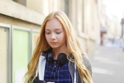Dziewczyna z blond włosami i słuchawkami na szyi przegląda telefon, zastanawiając się, jak zacząć rozmowę z chłopakiem.