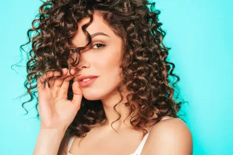 Mujer con cabello castaño y rizos definidos, tocando su cabello con la mano, sobre un fondo azul turquesa.