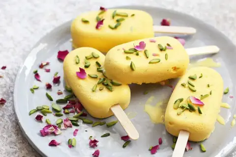 Quatre glaces jaunes, garnies de pistaches et de pétales de rose, sur une assiette. Un délicieux dessert iranien.