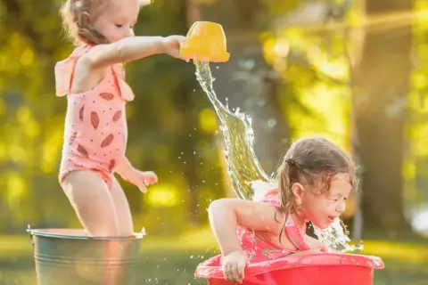 Bambine felici si divertono con giochi d'acqua sotto il sole estivo, spruzzandosi a vicenda con secchielli e bacinelle.