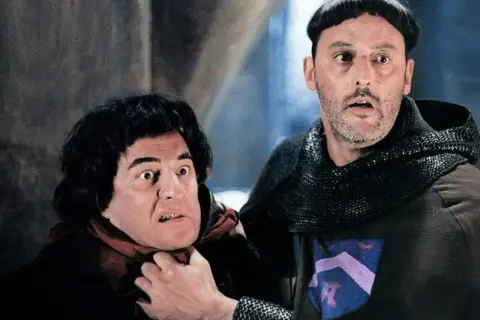 Goście, goście! Obsada filmu w średniowiecznych strojach, jeden z nich groźnie patrzy, drugi ma rękę na jego szyi.
