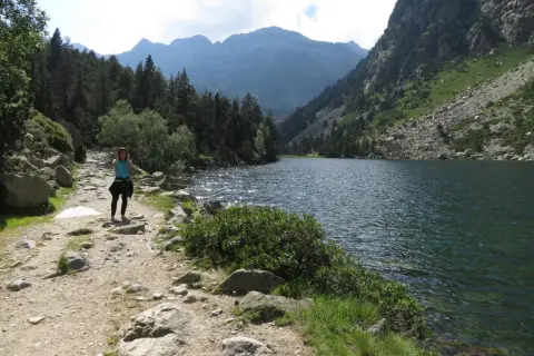 Una mujer camina por un sendero rocoso junto a un estany llong, rodeado de montañas y pinos.