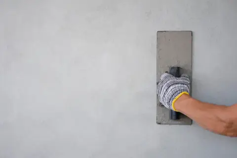 Mano con guanto che stende il rasante fino per interni su una parete grigia, creando una finitura liscia.