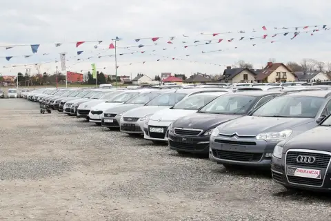 Rząd samochodów na placu, gotowych do sprzedaży. W tle domy i flagi. Możliwość zawarcia umowy komisowej samochodu.