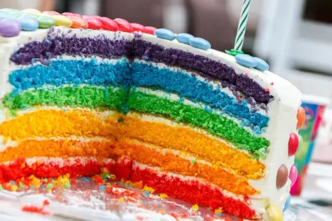 Kolorowy tort tęczowy z warstwami ciasta w odcieniach czerwieni, pomarańczu, żółci, zieleni, błękitu i fioletu, ozdobiony cukierkami.