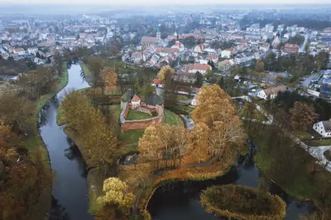 Zamek międzyrzecz otoczony jesiennymi drzewami i wodą, z miastem w tle.