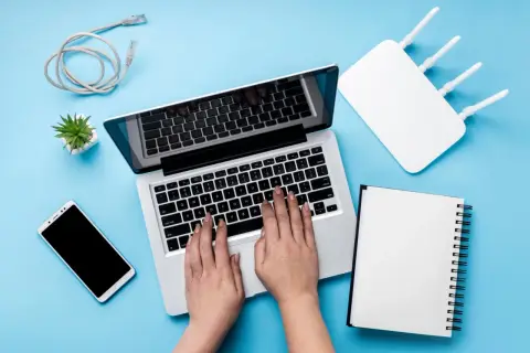 Ręce na klawiaturze laptopa, obok router wifi, telefon i kabel. Szukasz sposobu, jak zwiększyć zasięg wifi w laptopie?