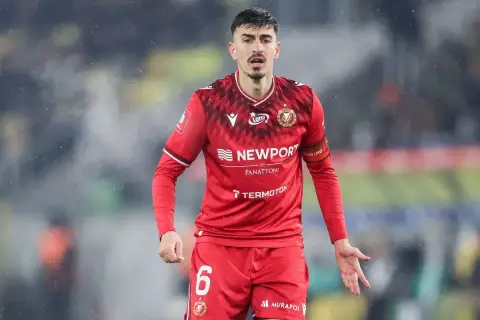 Piłkarz w czerwonej koszulce z numerem 6, z opaską na ramieniu z napisem "Widzew Łódź". W tle widać fragment stadionu, być może podczas meczu z Cracovią.
