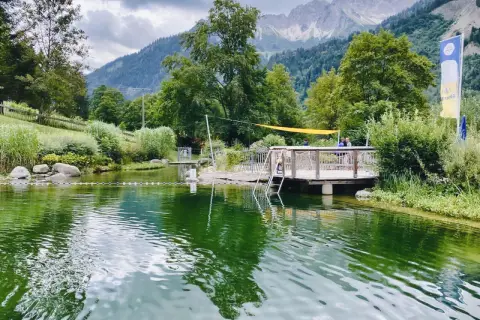 Ein idyllischer Badesee in Bad Hindelang, perfekt für das Wandern mit Kindern. Ein Steg mit Leiter lädt zum Sprung ins kühle Nass ein, während im Hintergrund majestätische Berge thronen.